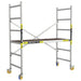 A silver aluminum mini scaffolding system with a wooden platform, yellow and black branded vertical sections, and caster wheels, shown on a plain white background.
